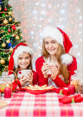 Merry Christmas and Happy Holidays. Mom with her daughter in red santa hats drinking tea with cookies at the Christmas tree