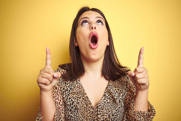 Young beautiful brunette woman wearing leopard shirt over yellow isolated background amazed and surprised looking up and pointing with fingers and raised arms.