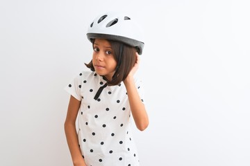 Beautiful child girl wearing security bike helmet standing over isolated white background smiling with hand over ear listening an hearing to rumor or gossip. Deafness concept.