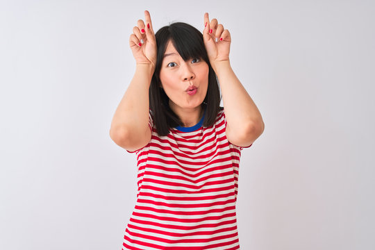 Young beautiful chinese woman wearing red striped t-shirt over isolated white background doing funny gesture with finger over head as bull horns