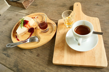 Cheesecake pie slice topped with strawberry syrup with spoon and fork , Espresso coffee in a white cup and a glass of honey on a wooden tray , Hot drink with brown table in background