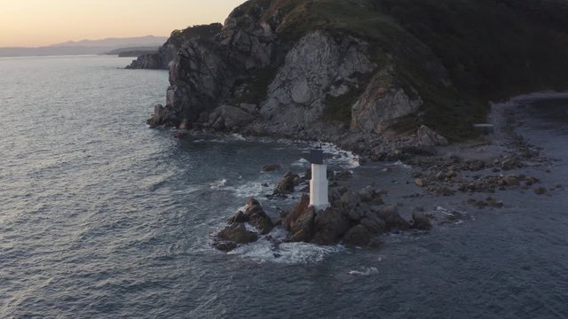 Slow lift up aerial shot of a small lighthouse and beacon, standing on the rocks near the big cliff with waves  crashing on them on the sunset. Russia, Far East.