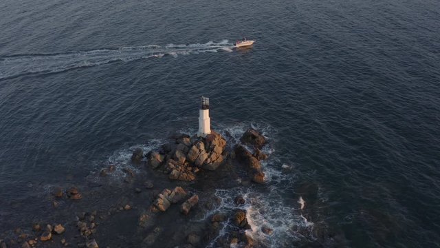 A small motor boat passing by a small lighthouse and beacon, standing on the rocks with waves  crashing on them on the sunset. Russia, Far East. Aerial shot.