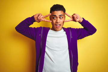 Fototapeta na wymiar Young brazilian man wearing purple sweatshirt standing over isolated yellow background Doing peace symbol with fingers over face, smiling cheerful showing victory