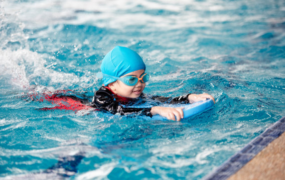 One girl wearing a swimsuit uses a foam pad to practice swimming in a swimming pool with a teacher. She happily and smiling.
