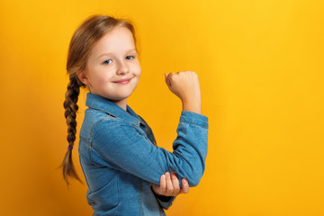 Closeup portrait of a little girl on a yellow background. Children's hand shows biceps. Girl power...