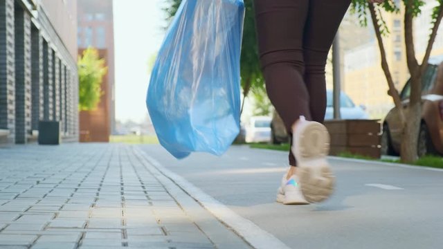 A housewife walks down the street with a bag of household garbage, bears throwing it away. The camera takes off her legs and follows her