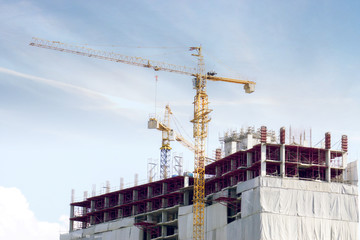 Building under construction with hoisting cranes working to move building materials under the blue sky and silken cloud.