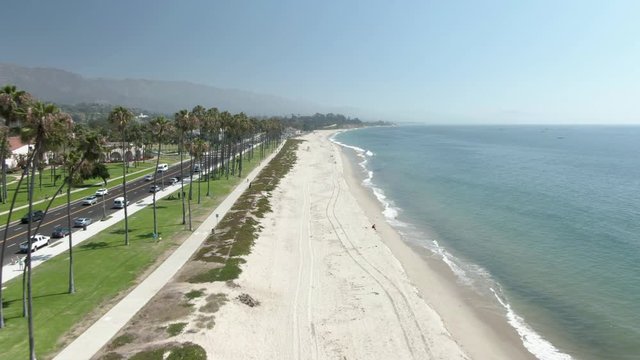 Low altitude aerial number 1 of an empty beach in Santa Barbara, California during a warm sunny day. Palm trees and public park on the left, Pacific ocean coastline on the right. Very few people.