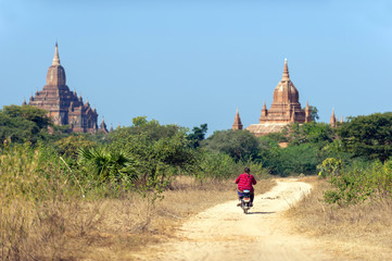 In the morning, clear sky, fat women wearing red shirts, motorbike on gravel road, behind a pagoda in Bagan, Myanmar.