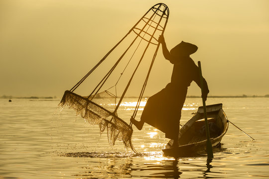 Closeup silhouette Burmese fisherman wearing a hat standing on the back of a boat using fishing equipment in the morning The mist faded at Inle Lake, Shan State in Myanmar.