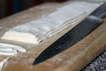 Rolled dough on wooden board prepeared for cutting homemade noodles with a large steel knife. Closeup, selective focus