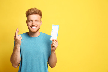 Portrait of hopeful young man with crossed fingers holding lottery ticket on yellow background,...