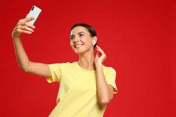 Happy young woman with wireless earphones taking selfie on red background