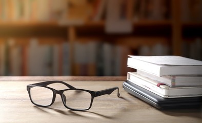 Eyeglasses with stacked books and black laptop on wooden table with flare light and blurred bookshelf in library  background