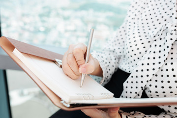 Close-up image of businesswoman sitting at office window and writing ideas in planner