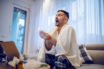 Ill caucasian employee in pajamas sitting on the sofa in living room covered with blanket and sneezing.