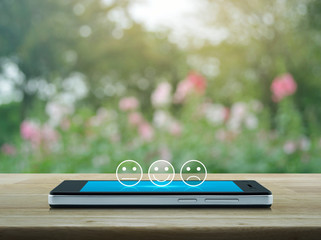 Excellent smiley face rating icon on modern smart mobile phone screen on wooden table over blur pink flower and tree in park, Business customer service evaluation and feedback rating online concept