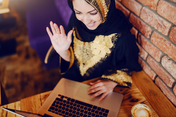 Top view of gorgeous smiling muslim woman dressed in traditional wear sitting in cafe, having video call and waving. On wooden table are laptop and coffee.