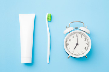 Alarm clock, toothbrush with green bristles and white tube of toothpaste on pastel blue background....