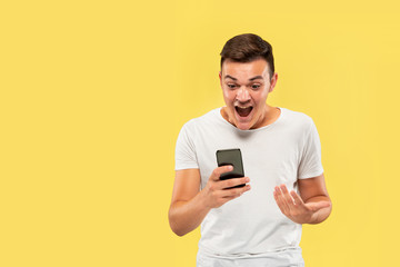 Caucasian young man's half-length portrait on yellow studio background. Beautiful male model in shirt. Concept of human emotions, facial expression, sales, ad. Using phone, looks crazy happy.