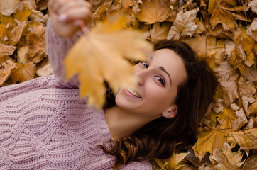 Happy young attractive woman with curly hair is standing in the park and covering her face with yellow autumn maple leaves. Cute girl enjoying warm weather. Autumn mood. Enjoy season