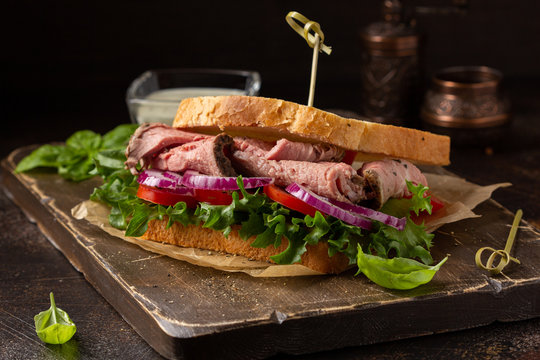 Roast beef sandwich with tomato, onion, lettuce and mustard sauce on dark background. Delicious healthy lunch with meat, hearty food