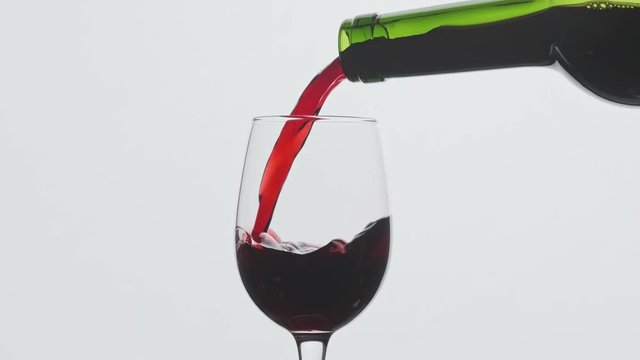 Red wine pouring in wine glass at white background. Close-up shot. Slow motion of pouring red wine from bottle into goblet. Red wine forms beautiful wave in 4k, UHD