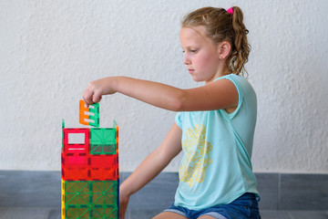 Beautiful teenage girl playing with lots of colorful plastic blocks constructor and builds house.