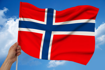 Fototapeta premium hand holds against the background of the sky with clouds the colored flag of Norway on the texture of the fabric, silk with waves, close-up