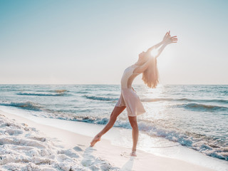 Slender beautiful ballerina in white dress dancing ballet on sea or ocean sandy beach in morning light. Concept of art, nature beauty - obrazy, fototapety, plakaty