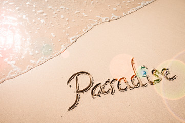 Paradise message handwritten on the smooth sand of an empty beach with lens flare over an incoming wave of copy space