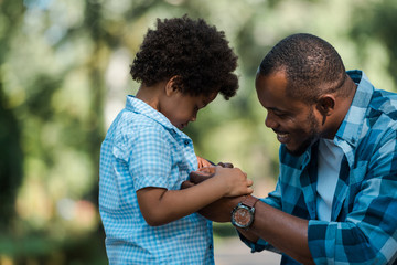side view of cheerful african american father holding hands with sad son