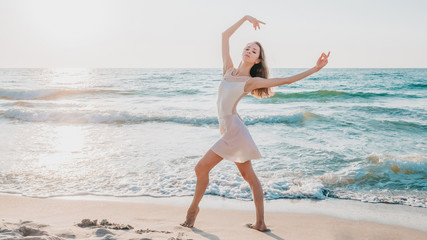 Slender beautiful ballerina in white dress dancing ballet on sea or ocean sandy beach in morning light. Concept of art, nature beauty