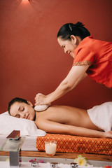 Obraz na płótnie Canvas side view of asian masseur doing back massage with herbal balls to woman in spa