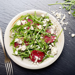 Top view at clay dish with beetroot arugula and feta cheese salad on slate stone tray closeup view. Fork aside