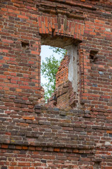 Old wall of old masonry of red brick with a dismantled window through which you can see the green foliage of the tree.