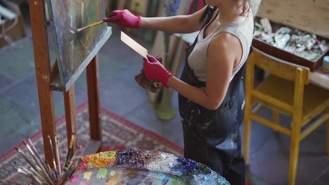 Unrecognizable female artist making painting reproduction on canvas. Woman working at easel holding picture with painting in her hand