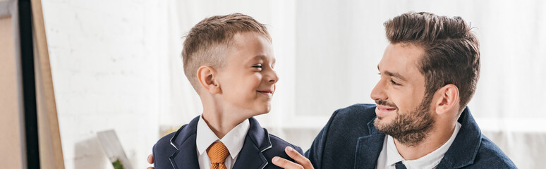 Fototapeta premium panoramic shot of smiling son and dad in formal wear looking at each other