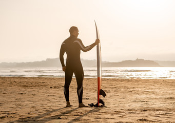 Rear view of strong surfer with surfboard on the beach at sunset or sunrise.