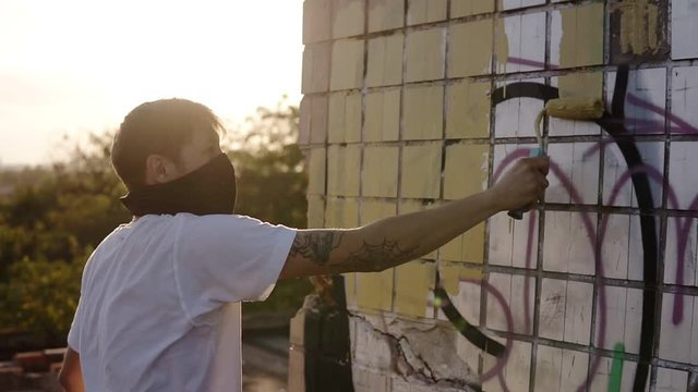 Caucasian guy is preparing the wall for graffiti, painting graffiti on abandoned building with roller paint into beige colour. Man is wearing casual clothes and black scarf on the face