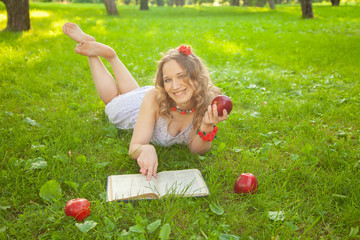 caucasian happy student girl in cute white dress rest on the green summer grass with book and red apples