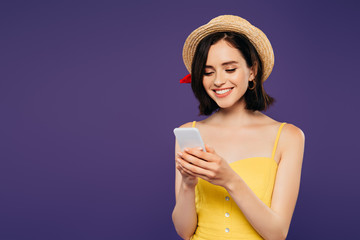 smiling girl in straw hat using smartphone isolated on purple