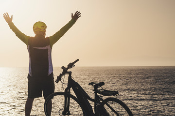 man in front of sea with arms up looking the horizont at the sunset with his bike - freedom...