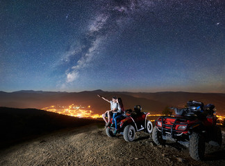 Happy couple man and woman trevelers sitting together on atv quad motorbike on the top of mountain. Man pointing at beautiful night sky full of stars, Milky way, luminous city on background