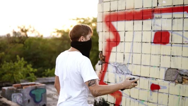 Young caucasian guy standing outdoors and trying his aerosol spaying it to the air then starting to paing the building wall. Daytime, sun shines on the background. Man wearing a white T shirt and