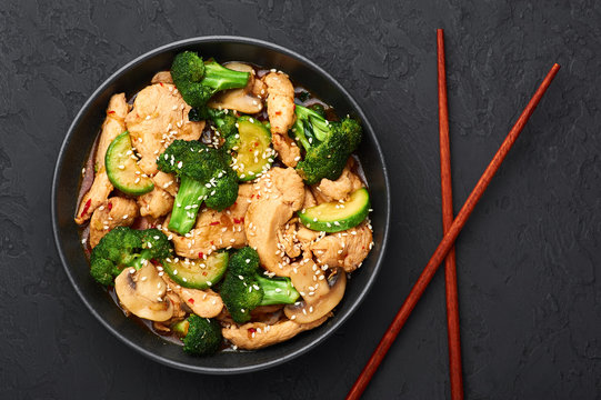 Hunan Chicken in black bowl at dark slate background. Hunan Chicken is chinese or indo-chinese cuisine takeaway dish with broccoli, zucchini, shiitake mushrooms and hunan sauce. Top view