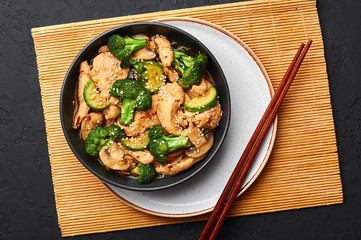 Hunan Chicken in black bowl at dark slate background. Hunan Chicken is chinese or indo-chinese cuisine takeaway dish with broccoli, zucchini, shiitake mushrooms and hunan sauce. top view