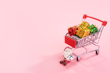 Christmas shopping concept, mini red shop cart trolley with Santa Claus toy and gift box isolated on pale pink background, blank copy space, close up