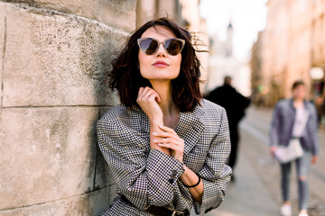Beautiful brunette young woman wearing jacket and black sunglasses walking on the street, posing...